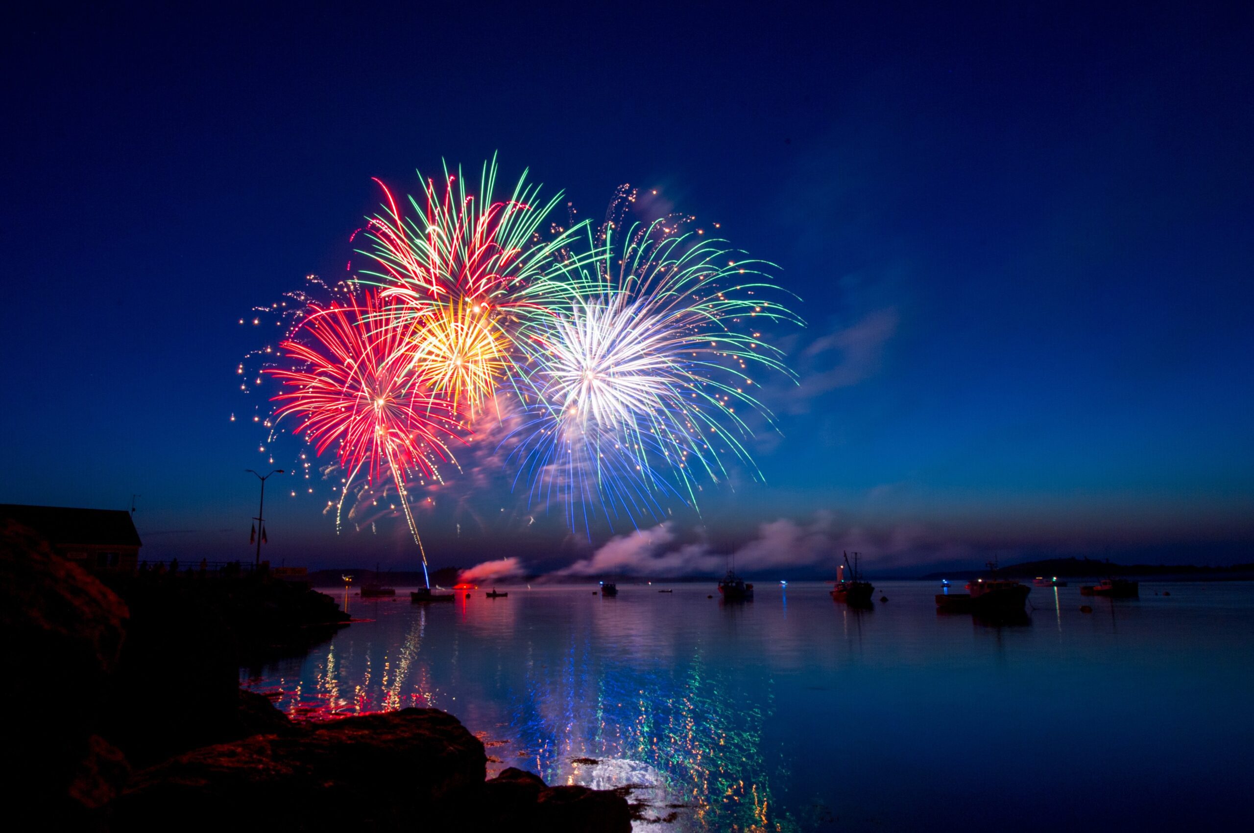 Fireworks explode in rainbow colors in a dusk-filled sky. The fireworks have been set off, and reflect off of, a body of water.